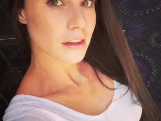 Hazel_Blaze poses with a close-up view of her face and long dark hair, wearing a white scoop-neck top.