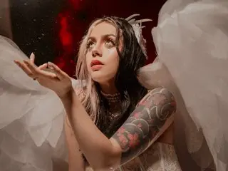 Hela_VXX poses with her hands near her face, showing her dark hair, heavy eye makeup, and large white wings against a dark red background.