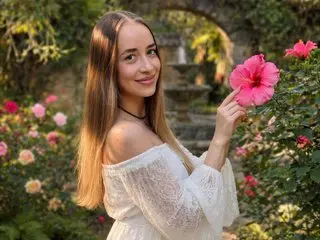 Smiling young woman in a white lace off-the-shoulder dress holding a pink hibiscus in a sunny garden.