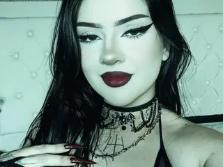 Gothic woman with dark makeup and lace choker posing in a moody bedroom setting.