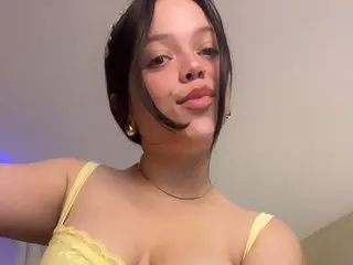 Helenna59 poses close up, wearing a yellow lace bra and showing her face, hair, and cleavage against a plain wall.