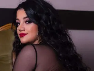 HellennaSweett poses with long wavy black hair, wearing a black sheer mesh top and red lipstick against a dark background.