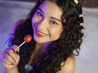 Smiling young woman with curly hair holding a red lollipop in a dreamy, purple-lit setting.