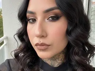 HeraM poses close-up, showing her face, dark wavy hair, and a tattoo on her neck.