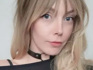 Heriath23 looks at the camera with blonde hair and a black choker, wearing a black top against a plain white background.