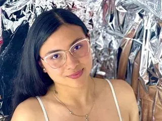 HimariSaenz poses with long dark hair and glasses, wearing a white strap top against a shiny silver foil background.