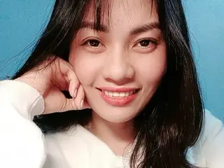 Hinna_x smiles while resting her chin on her hand, showing her long dark hair and face against a blue background.