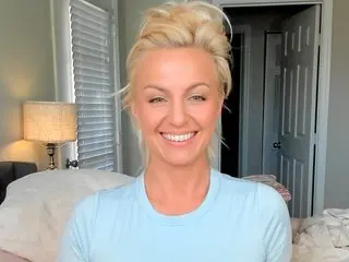 HollyKennedy smiles while wearing a light blue t-shirt, showing her blonde hair and face in a bright indoor setting.