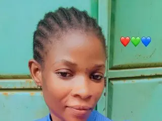 Honiegal looks directly at the camera with her dark hair pulled back in a braided style, showing her face and shoulders against a plain green background.