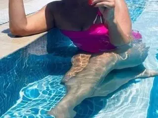 HornyHarriet_XX poses nakedly in a swimming pool, showing her bare breasts and stomach while leaning against the blue tiled edge.