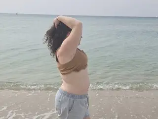 Curvy woman in a brown bikini top and denim shorts relaxing on a sunny beach by the ocean.