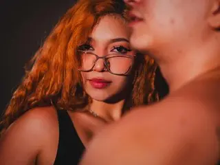 Hottie67 poses close-up with long red hair and glasses, showing her face and bare shoulders against a dark background.