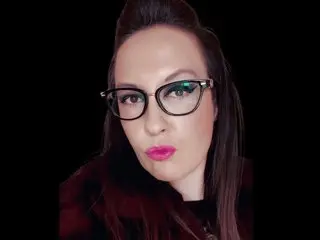 Humiliatrixxxx looks into the camera wearing black-rimmed glasses, with her dark hair pulled back and pink lipstick, set against a black background.