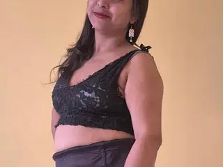 IamMeera poses in a black lace crop top and dark shorts against a plain yellow wall.
