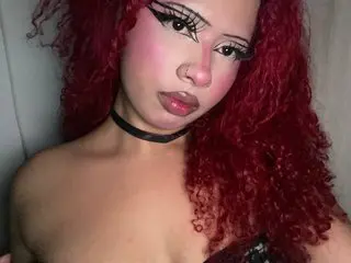 IdunBrunette poses with her long red hair, wearing a black choker and a black lace top, showing her bare shoulders and chest.