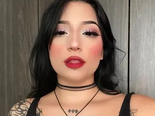 ImEmilyy444 poses with long dark hair and red lipstick, wearing a black choker and a black tank top against a grey wood wall.