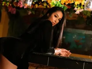 ImLau poses in a black long-sleeve top with her long dark hair down, leaning over a dark surface against a colorful floral background.