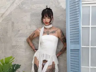 Im_Doll_Bella poses in a white halter top and shorts, showing off her tattoos against a stone wall.
