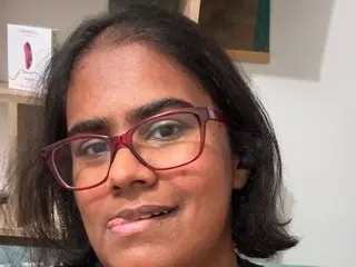 Indiangodddessss looks at the camera with a slight smile, showing her dark hair and red-framed glasses in a close-up headshot.