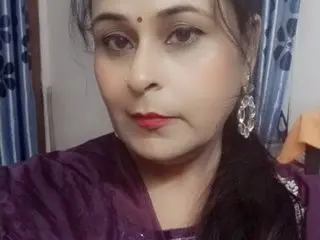 Indianpyari looks at the camera with long dark hair and red lipstick, wearing a purple top against a soft indoor background.