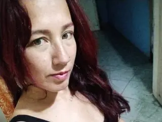 IriisRoses shows her face and shoulders with long red hair, looking directly at the camera in a close-up shot.