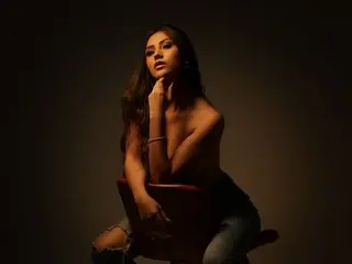 IsabellaSaenz poses shirtless, looking at the camera with her hand touching her neck against a dark background.