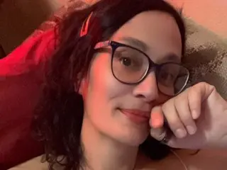 IsabellaSarai looks at the camera with a slight smile, wearing black-rimmed glasses and showing her dark hair in a close-up shot.