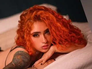 IsabellaVillalobos has long, wavy orange hair and looks directly at the camera while lying on a white bedsheet.