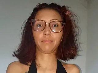 Portrait of a woman with reddish hair and glasses wearing a black halter top against a plain wall.