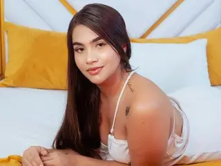 IsisFerrer with long dark hair poses in a white top against a yellow and white bed setting, showing her bare shoulders and collarbone.