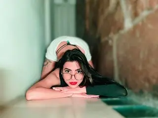 IsisRoosexx poses with her long dark hair and glasses, wearing a white skirt and a black top while leaning forward on a flat surface.