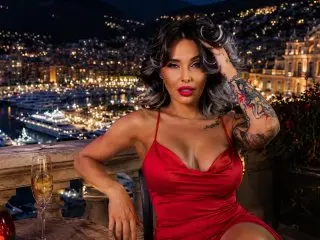 IssaSmithx poses with her hands in her dark wavy hair, wearing a red low-cut top while sitting outdoors against a blurred city night view.