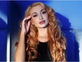 IvaRoberts poses with her long, wavy blonde hair, wearing a black top against a blue background.
