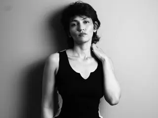 Moody black and white portrait of a woman in a black tank top leaning against a plain wall.