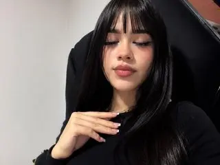 IvonJanice with long black hair and bangs poses in a black top against a dark chair.