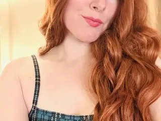 Smiling redhead woman with wavy hair wearing a blue plaid tank top in a bright indoor setting.