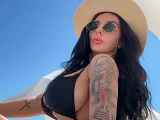 IvyWiksen poses in a tan hat and black bikini top, showing her long dark hair and shoulder tattoo against a clear blue sky.
