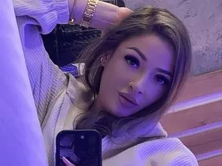 Izza_Bella poses with a hand behind her head, showing her face, long brown hair, and a light-colored striped top or sweater.
