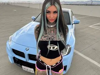 IzzyKreame poses in a black tank top and black undies, showing off her long dark hair and tattoos while leaning against a light blue car outdoors.