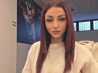 JANNYIE poses with long red hair, wearing a white v-neck sweater against a dark background.