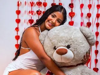 JANYLOVE with long dark hair smiles while hugging a large grey teddy bear against a background of red heart decorations.