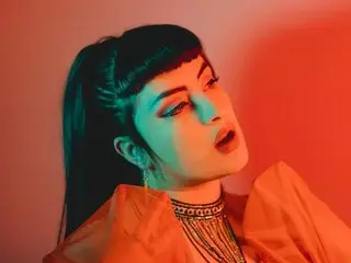 JESSICABUNNY_69 poses with her dark hair in a high ponytail and blunt bangs, wearing an orange top against a red and pink background.