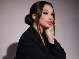 JOSEPHIYN poses with her hand near her face, wearing a black blazer against a plain grey background.