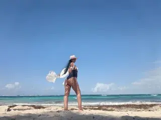 Stylish woman in a black swimsuit and sun hat walking on a sunny tropical beach under a clear blue sky.