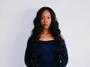 JadeCams poses with her long, wavy black hair down, wearing a dark blue off-the-shoulder top against a plain light background.