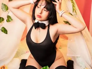 Jade_Rous poses with her hands behind her head, wearing a black bodysuit and white collar with a black bow, showing her cleavage and black hair.