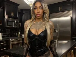 Januaryvice poses in a black corset and black lace gloves, showing off her long blonde hair and cleavage against a dark kitchen background.