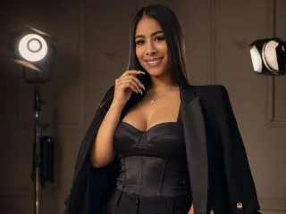 JasminKoch smiles in a black corset and blazer while posing in a studio with bright lights.
