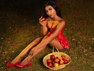 JasminMale sits on the grass wearing a red silk slip, holding a red apple near her chest with her legs bent and exposed.