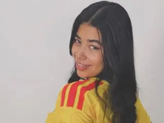 Jasmin_606 smiles over her shoulder, wearing a yellow track jacket with red stripes against a plain white background.